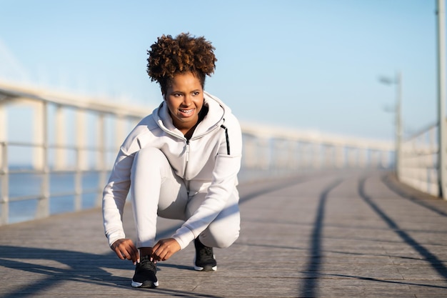 Улыбающаяся черная женщина завязывает шнурки на кроссовках перед пробежкой на свежем воздухе