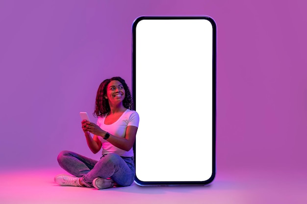 Smiling black woman using mobile phone while sitting near huge blank smartphone