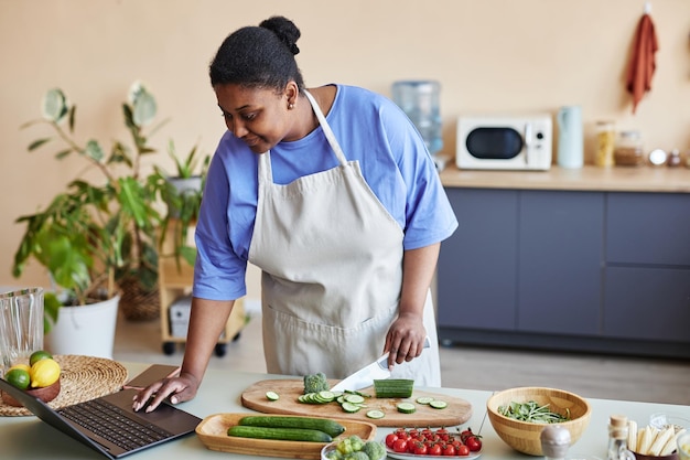 Photo smiling black woman wearing apron in kitchen and cutting vegetables