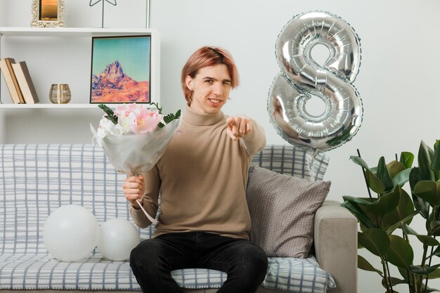 Smiling blinked points at front handsome guy on happy women day holding bouquet sitting on sofa in living room