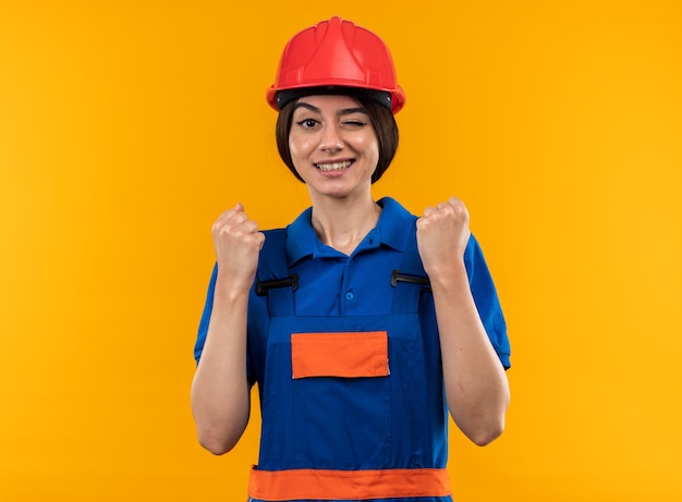 Smiling blinked young builder woman in uniform showing yes gesture isolated on yellow wall