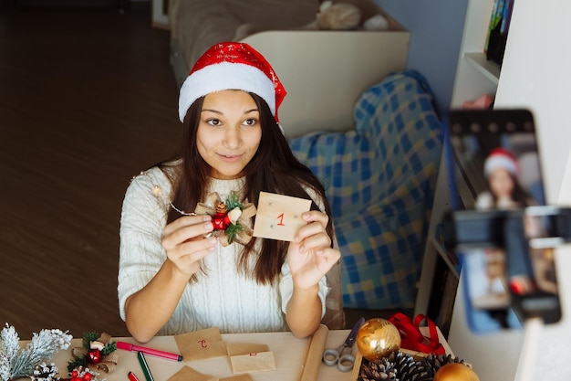 クリスマスの帽子をかぶった笑顔のブロガーの女の子がアドベントカレンダーを作成し、彼女の携帯電話で写真を撮ります