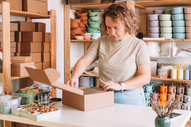 Smiling blond curly woman packing and wrapping shipment silicone baby dishes in cardboard delivery box