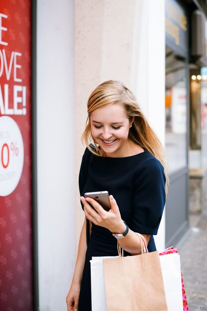 smiling blond girl in black dress looking to a mobile phone and carrying bags after shopping