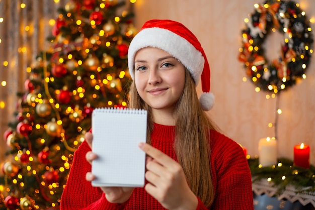 Smiling blond girl in red sweater and Santa hat pointing to note book Christmas concept