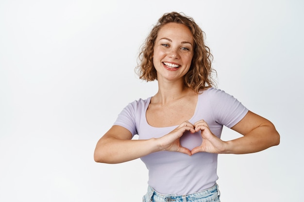 Smiling blond girl shows heart gesture, showing love and care for someone, standing on white