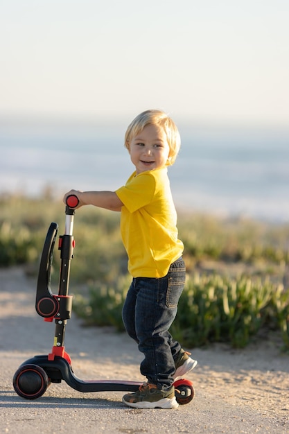 Smiling blond little boy stands on the road holding by scooter