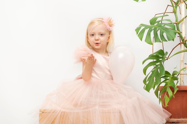 Smiling Blond little girl with balloon Little girl in pink dress and crown hoop on headMonstera flower on background