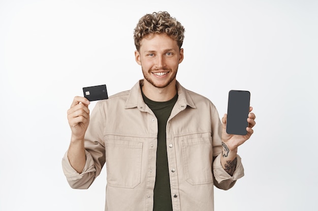 Smiling blond man showing smartphone screen and credit card standing in casual clothes Cellular technology concept
