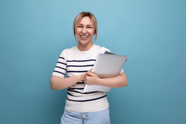 Sorridente studentessa bionda in abbigliamento casual con un computer portatile in mano su sfondo blu