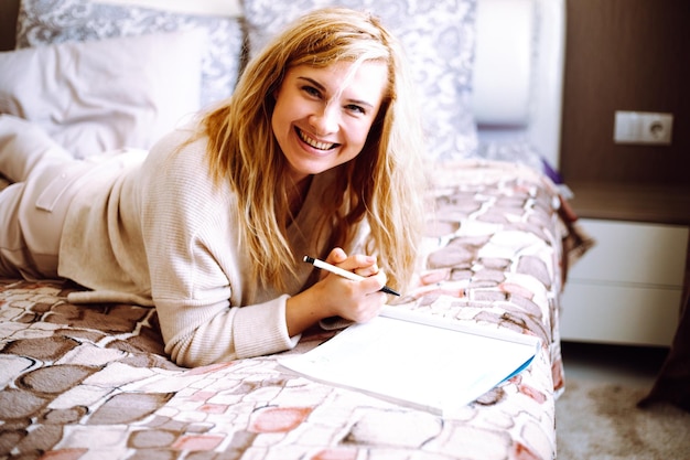 Улыбающаяся блондинка, лежащая в постели дома, смотрит в камеру, пишет в учебнике, изучает бизнес или учится