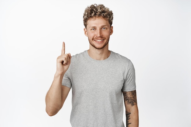 Smiling blond young man showing sale banner pointing finger up and looking at camera standing over white background