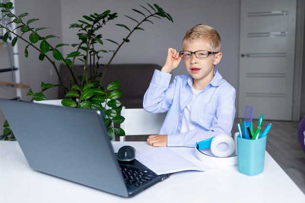 Улыбающийся белокурый мальчик в очках делает видеозвонок через ноутбук во время дистанционного обучения дома