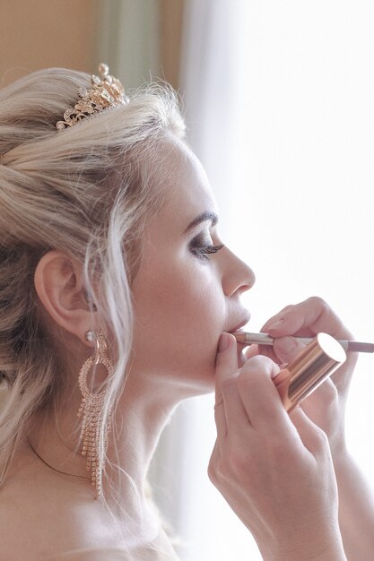 smiling blonde bride in a hotel room paints her lips