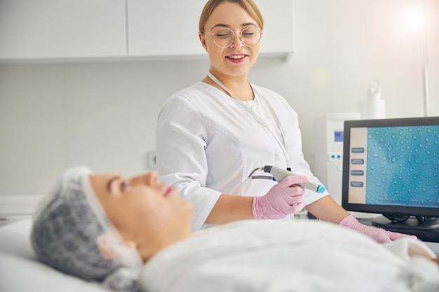 Smiling blonde female dermatologist looking at her calm young dark-haired Caucasian patient in a bathrobe