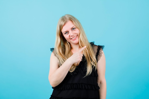 Smiling blonde girl is pointing right with forefinger on blue background