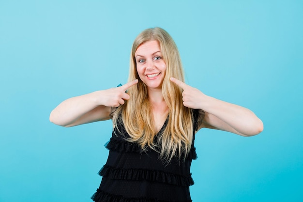 Smiling blonde girl is showing her smile with forefingers on blue background