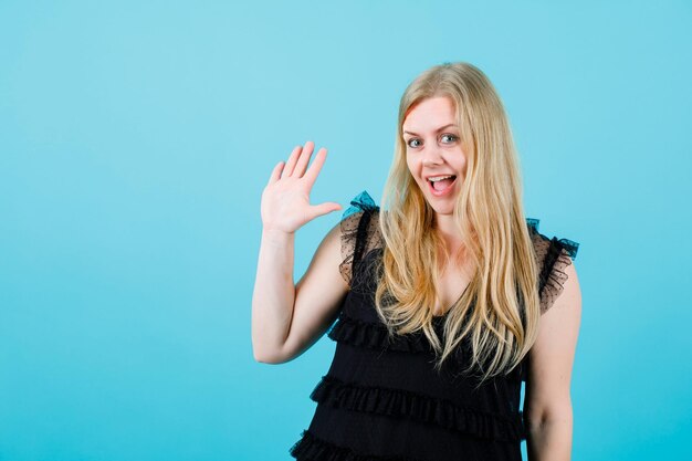 Smiling blonde girl is showing hi gesture by raising up her handful on blue background