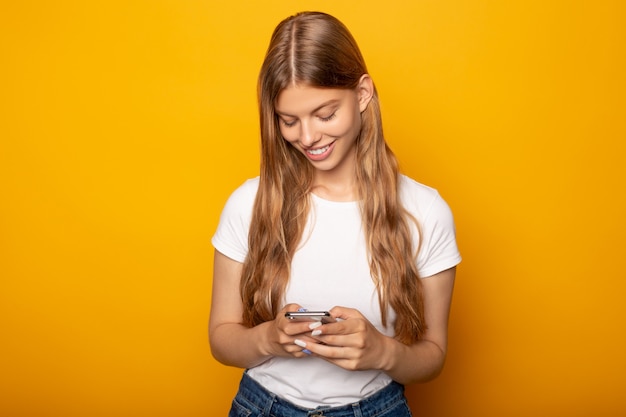 smiling blonde girl using smartphone isolated on yellow