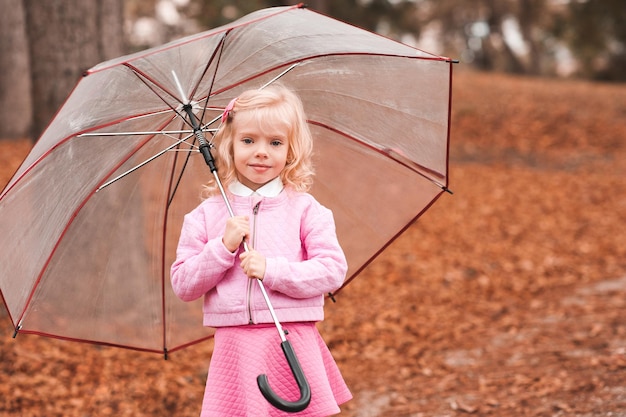 Smiling blonde kid girl holding umbrella standing in autumn park