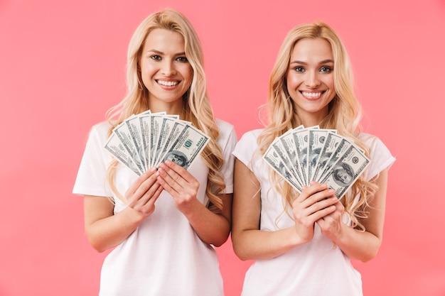 Smiling blonde twins wearing in t-shirts holding money and looking at the front over pink wall