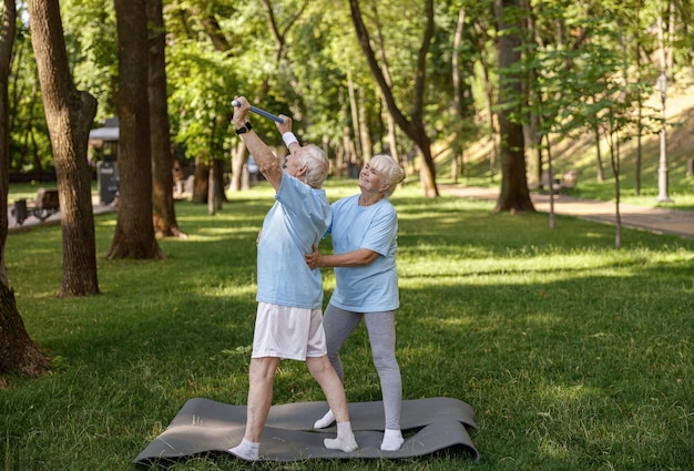 Smiling blonde wife helps senior man to do sports exercises with bar in green park