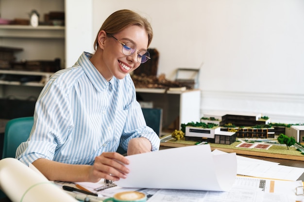 smiling blonde woman architect in eyeglasses working with drawings while designing draft at workplace