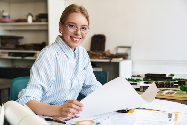 улыбающийся белокурая женщина-архитектор в очках, работающая с чертежами при разработке проекта на рабочем месте