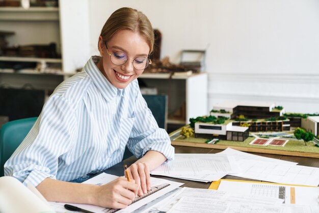 smiling blonde woman architect in eyeglasses working with drawings while designing draft at workplace