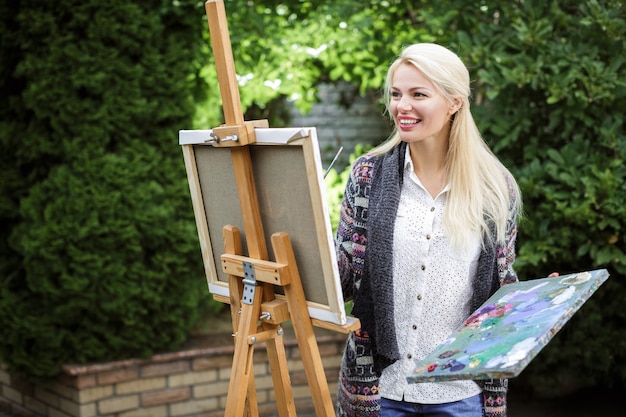 Smiling blonde woman artist with a brush in her hand draws on canvas in the nature.