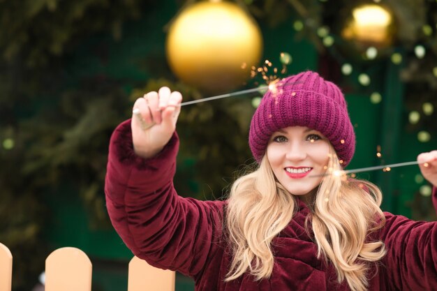 Smiling  blonde woman dressed in red knitted hat and warm coat holding glowing sparklers at the Christmas tree