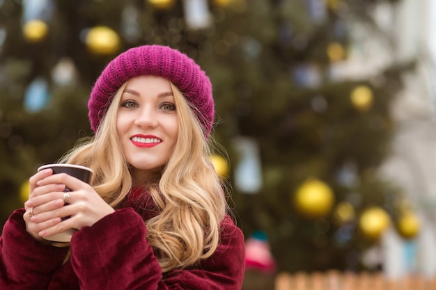 Donna bionda sorridente vestita con abiti invernali e che beve caffè all'albero di natale