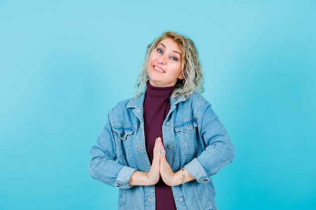 Smiling blonde woman is looking at camera by holding hands together on blue background