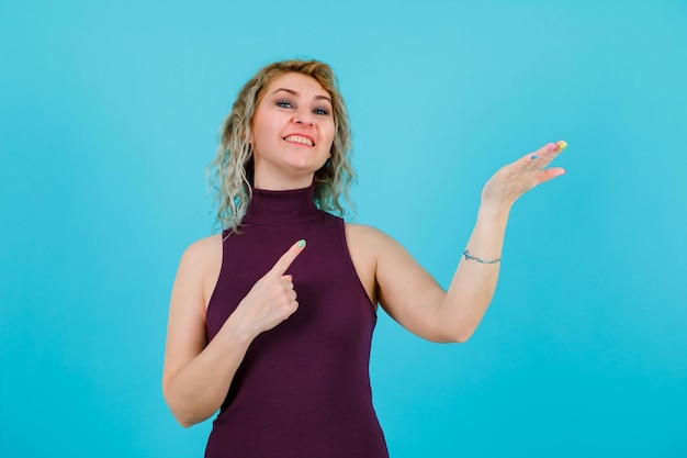 Smiling blonde woman is raising up her hand ful and pointing it with forefinger on blue background