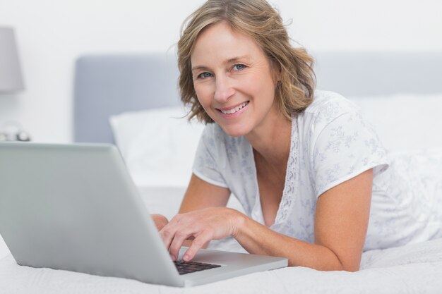 Donna bionda sorridente che si trova sul letto facendo uso del computer portatile