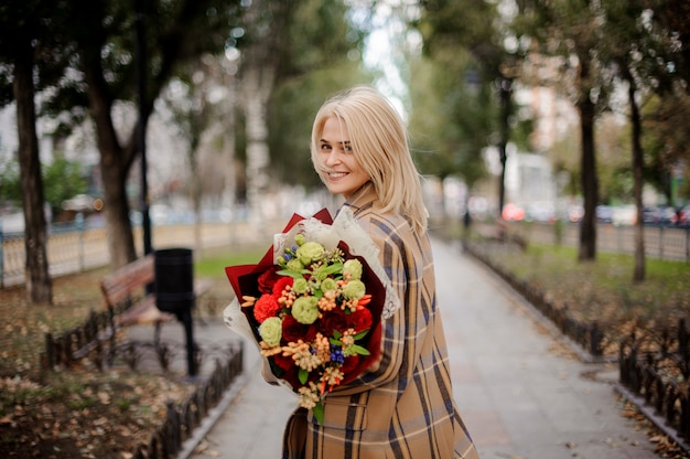 Donna bionda sorridente in cappotto scozzese che tiene un mazzo luminoso di fiori che cammina sul vicolo