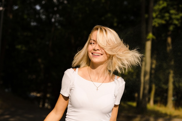 Улыбающаяся блондинка в белой футболке с волосами в движении позирует в парке