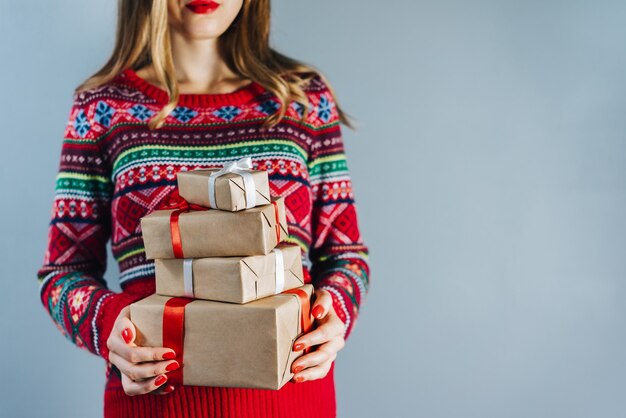 smiling blonde woman with red lips and polished nails holding bunch of gift boxes wrapped in craft paper and decorated with red satin ribbon. Christmas concept