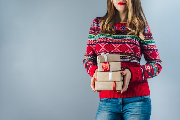 クラフト紙に包まれ、赤いサテンのリボンで飾られたギフトボックスの束を保持している赤い唇と磨かれた爪を持つ笑顔のブロンドの女性。クリスマスのコンセプト
