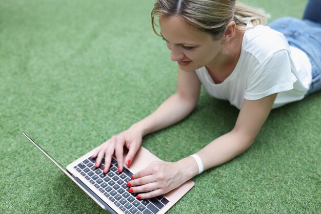 Smiling blonde woman working on laptop lay on grass remote job female freelancer outdoors