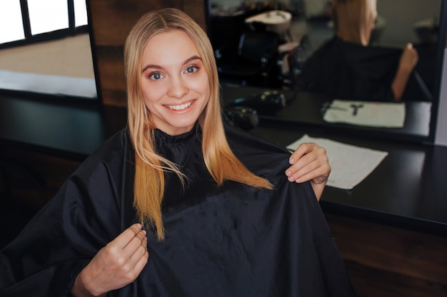 Giovane donna bionda sorridente con mantello nero che si siede nella sedia dei parrucchieri davanti allo specchio mentre aspettando nuovo haitcut in salone. bellezza e persone