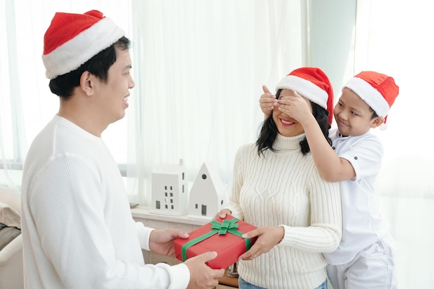 Ragazzo sorridente che copre gli occhi della madre quando papà le fa il regalo di natale