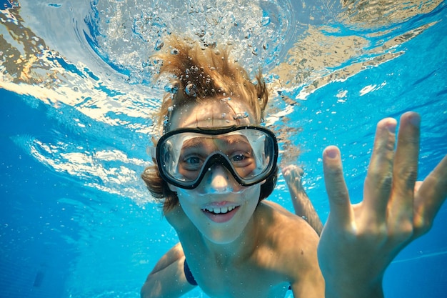 Улыбающийся мальчик в очках для дайвинга плавает в чистой прозрачной воде в бассейне и смотрит в камеру