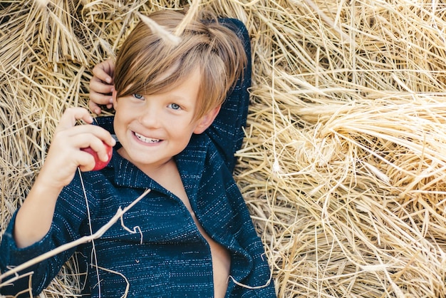 A smiling boy holds a bitten apple with one hand Happy childhood Fairhaired boy lies on hay backgrou