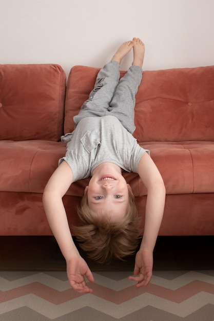 笑顔の少年は逆さまにソファに横たわって、カメラを見てソファで休んでいるかわいい幼児