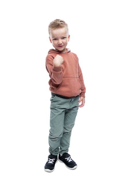 Улыбающийся мальчик стоит Симпатичный блондин с модной прической в коричневой толстовке с капюшоном и серых брюках Радость и позитив В полный рост Изолированный на белом фоне Вертикальный