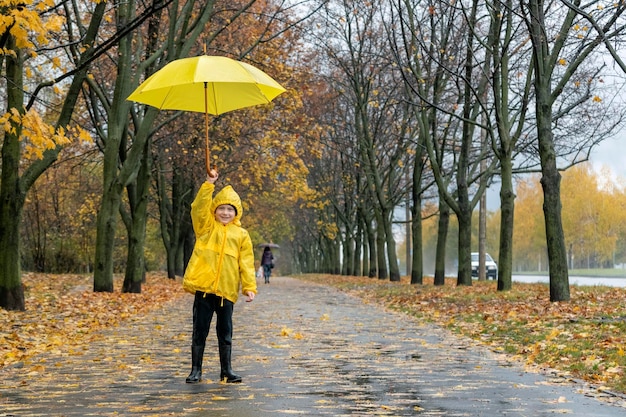 笑顔の少年が黄色い傘を持って公園で遊ぶ 秋の雨の通りの子供が歩いている