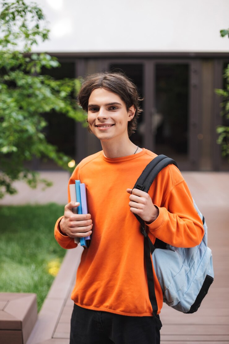Smiling boy standing with backpack and books in hand and happily in courtyard of university