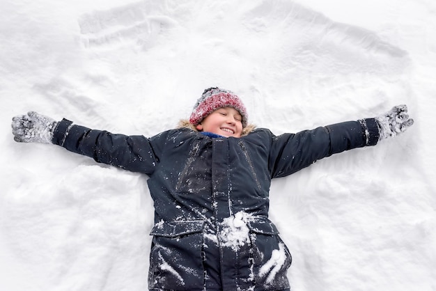 暖かい服を着た笑顔の少年が仰向けになって雪の中で天使を見せている