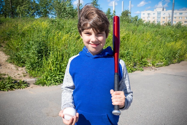 Smiling boy with baseball bat and ball in blue hoodie against green grass outdoors on sunny day